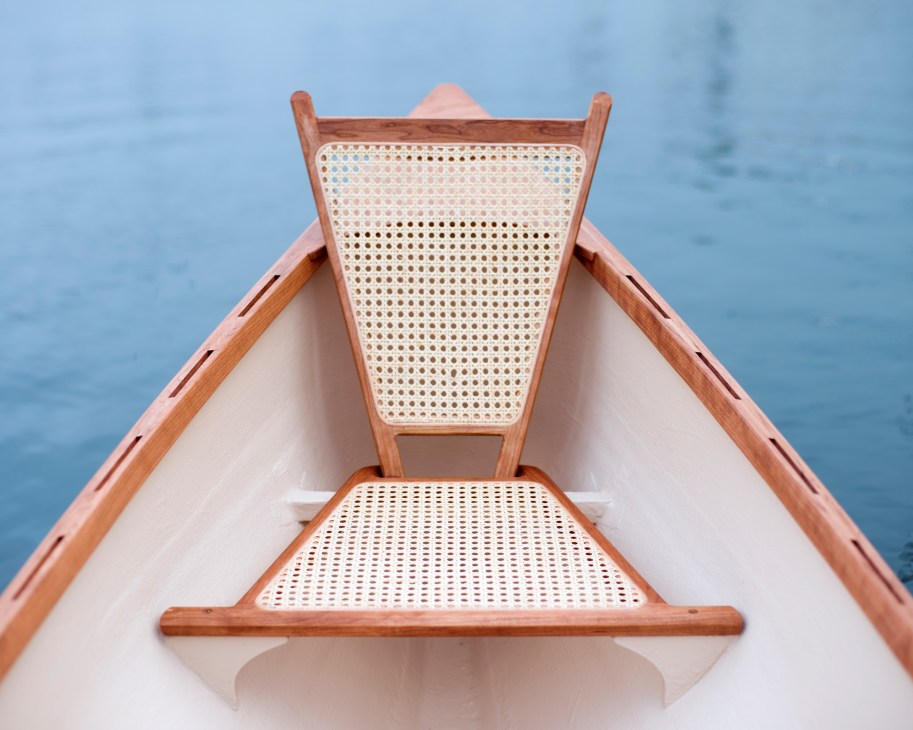 Interior view of a wooden rowboat with woven seats and backrest, looking toward the bow on calm blue water.