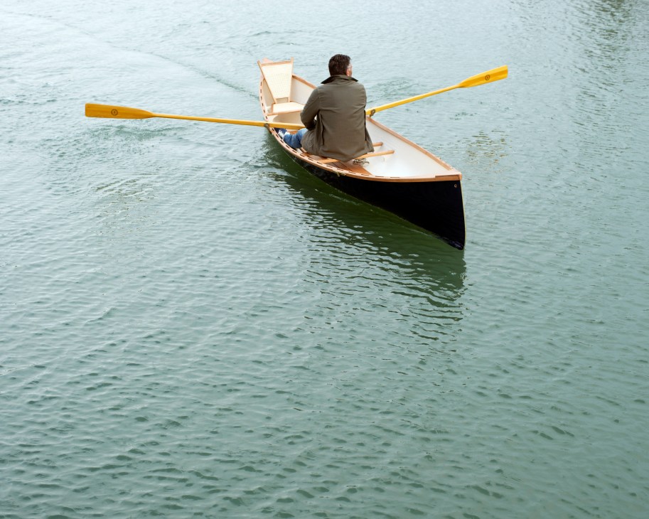 Person rowing a wooden rowboat with yellow oars on calm water.