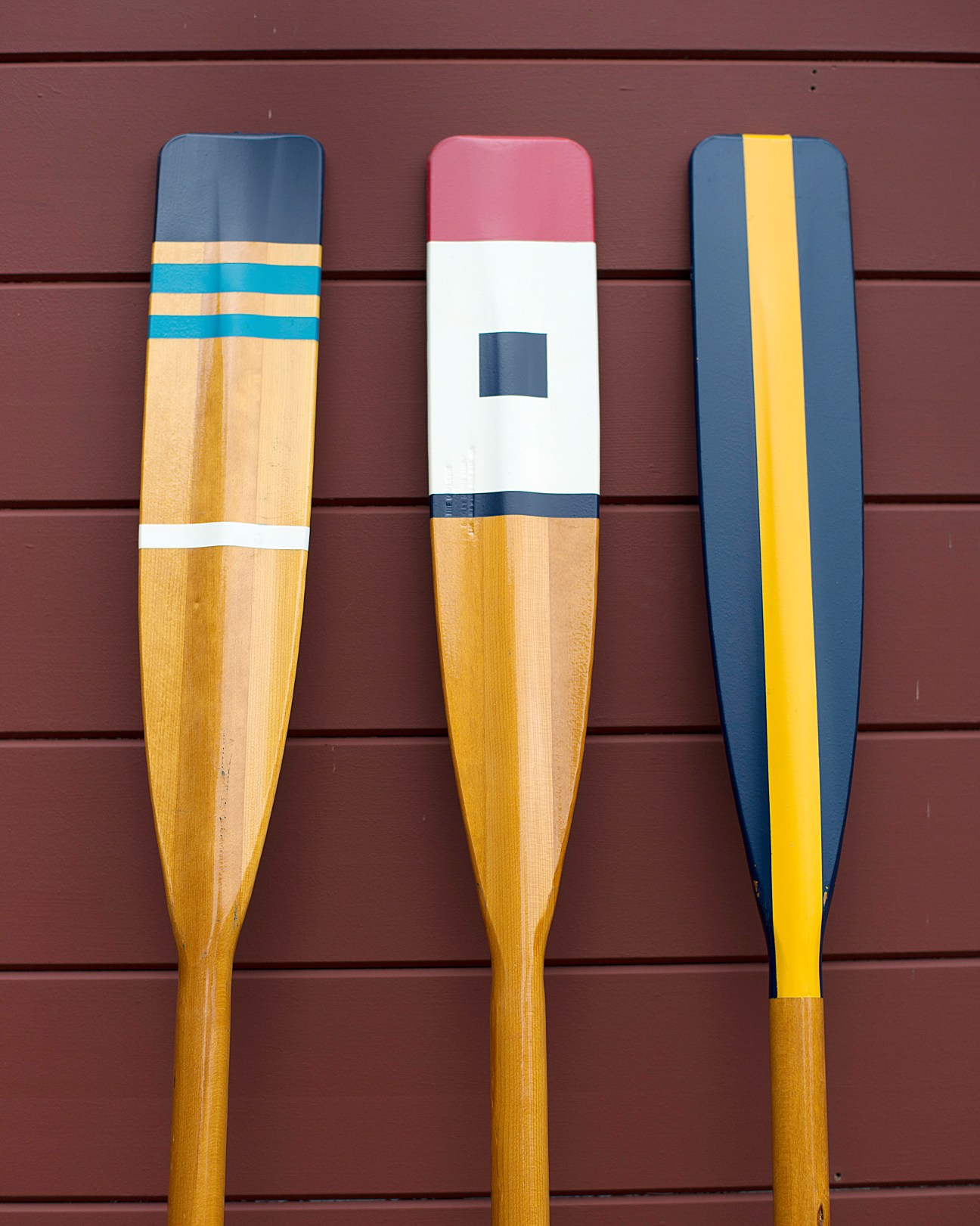 Three wooden oars with colored handles displayed against a maroon wall.