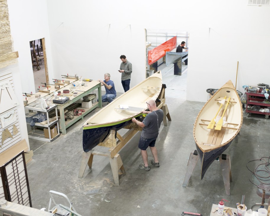 Interior view of Guideboat Co's workshop with workers and wooden rowboats in various stages of construction.