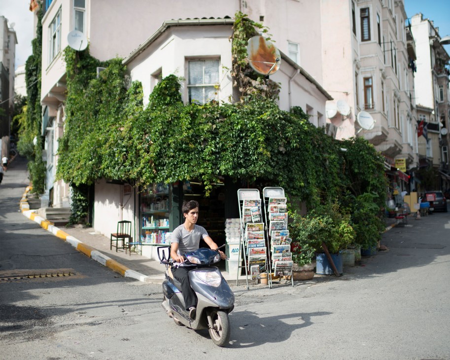 Man on scooter passes ivy-covered corner shop with newspaper racks in Istanbul's Beyoglu neighborhood.