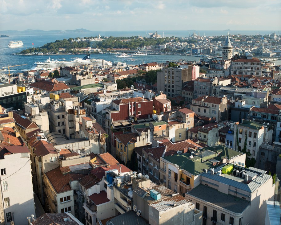 Aerial view of Istanbul showing densely packed buildings with red-tiled roofs, the Golden Horn waterway, cruise ships, mosques, and the Bosphorus strait in the distance.