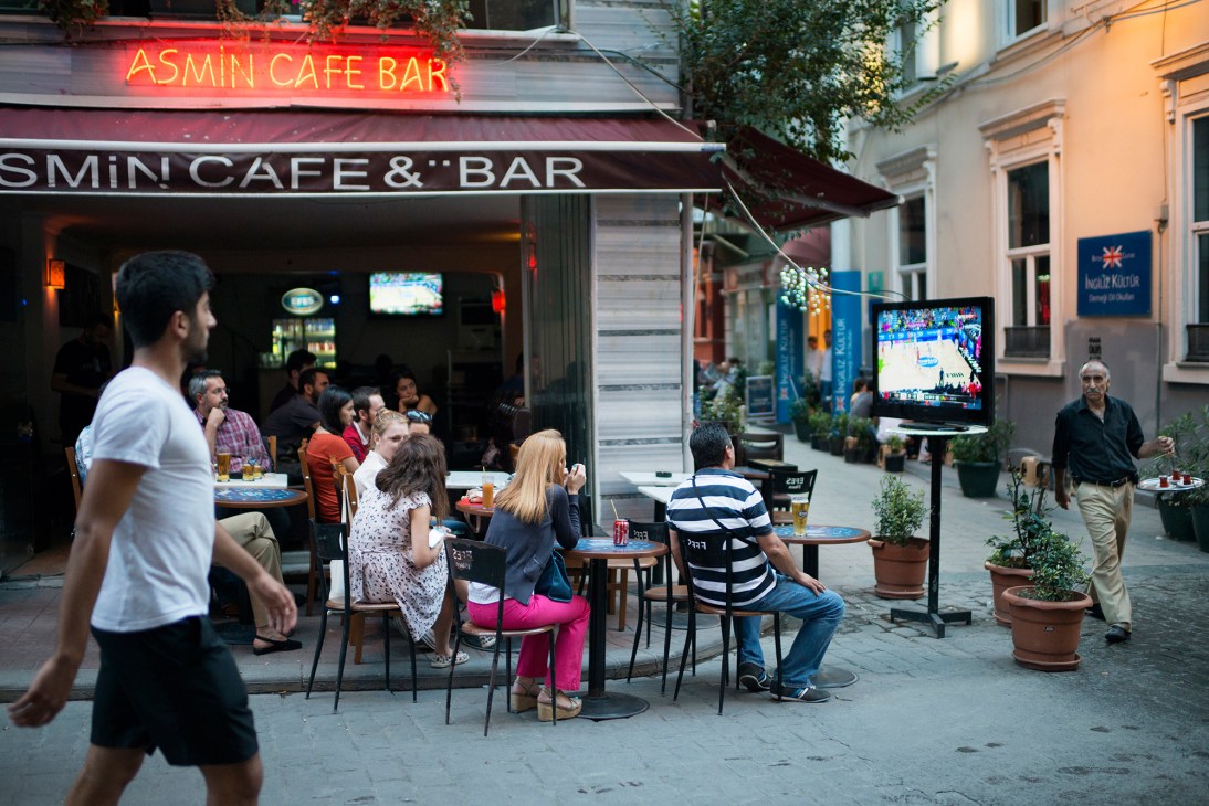 People sitting at outdoor café tables watching basketball on a TV screen near Taksim Square in Istanbul.