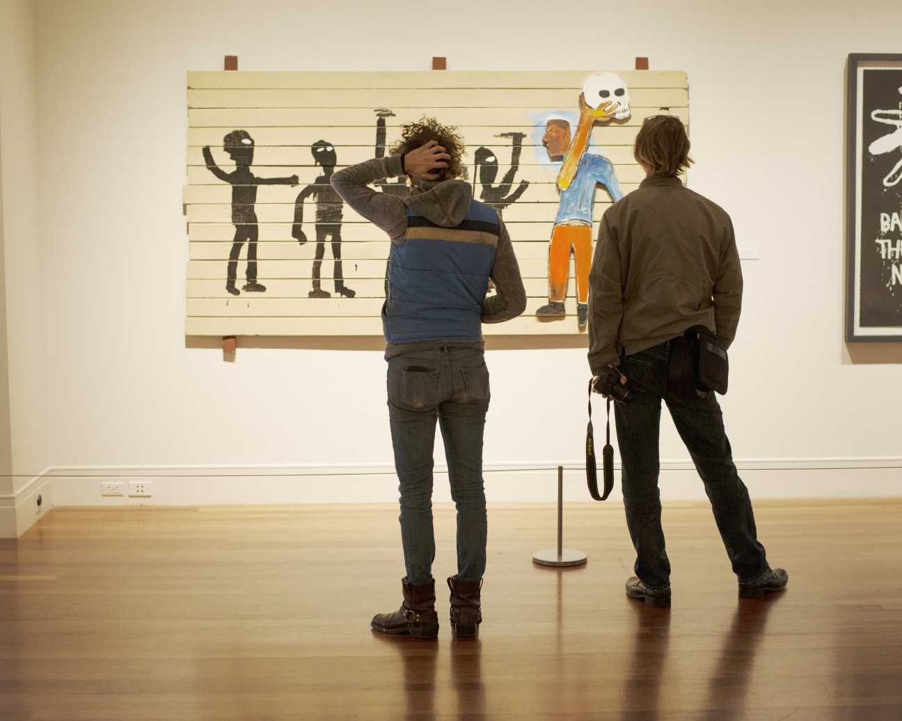 Two visitors viewing a Jean-Michel Basquiat artwork featuring black silhouettes and colorful figures on cream-colored wood panels at the Ogden Museum of Southern Art.