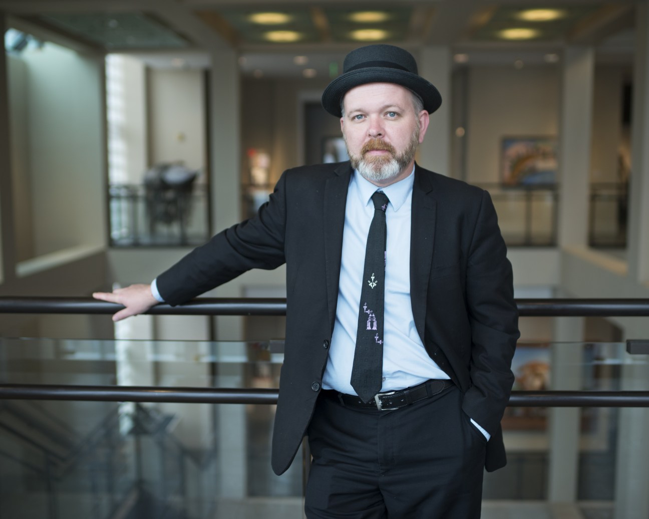 Bradley Sumrall, chief curator at the Ogden Museum of Southern Art, poses in a museum hallway wearing a dark suit, light blue shirt, and decorative tie with a black bowler hat.
