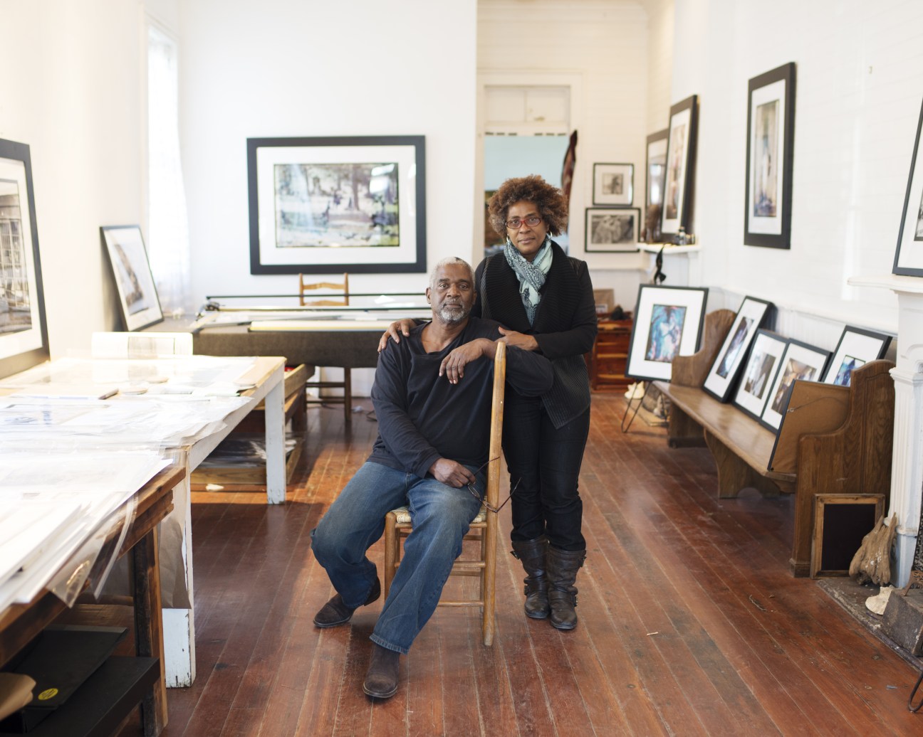 Two people pose together in an art gallery surrounded by framed artworks and display tables.