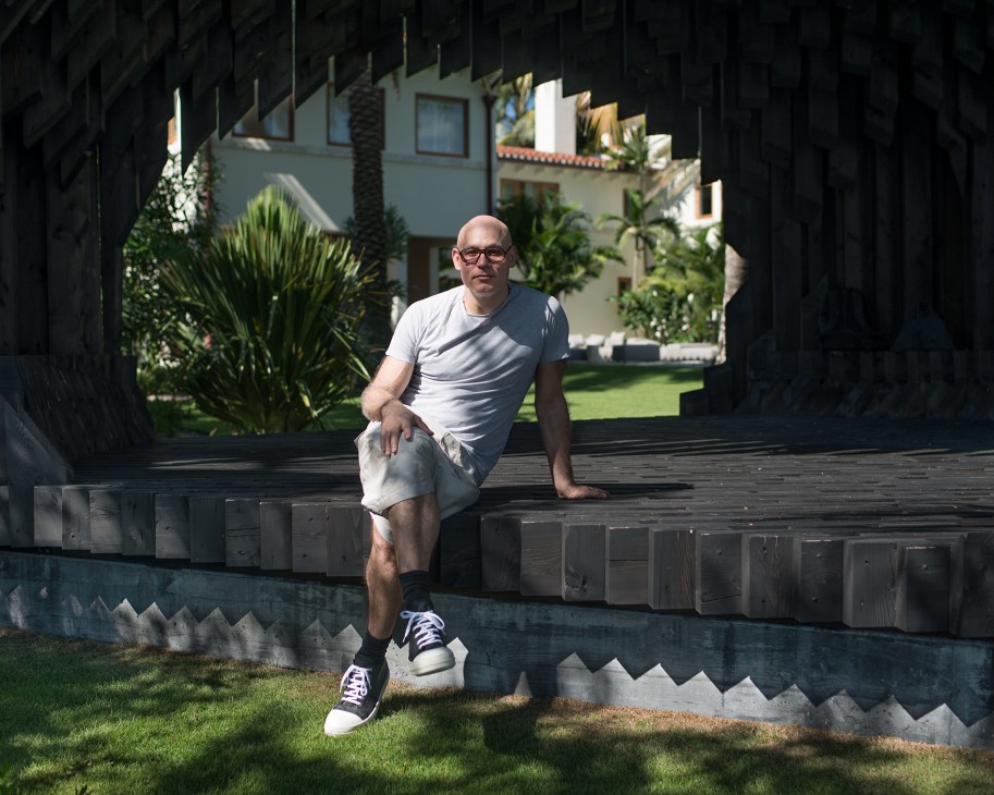 Craig Robins sits on a wooden bench in his Miami garden pavilion, with tropical plants and art deco architecture visible behind him.