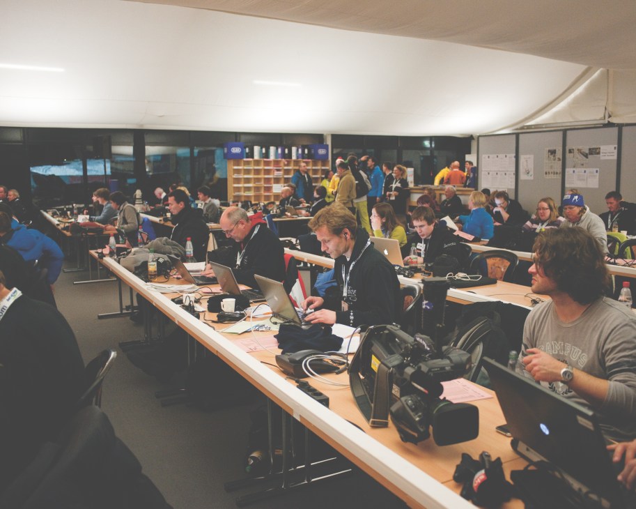 Journalists working at long tables in a press center with laptops and notebooks during a ski jumping event.