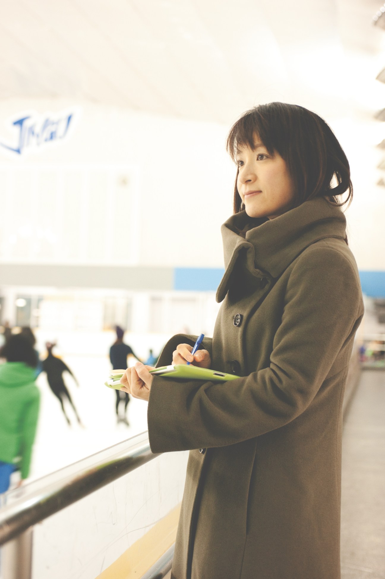 Yoshie Noguchi takes notes at an ice rink while figure skaters practice in the background.