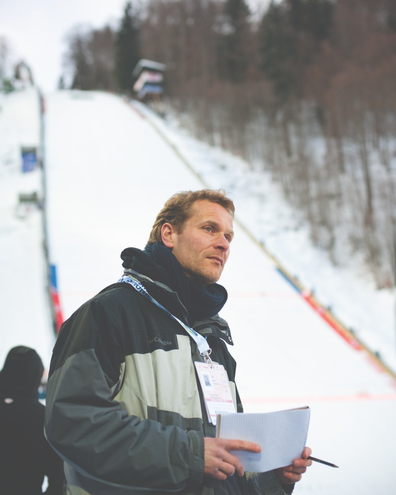 Thomas Hahn at a ski-jumping competition in Oberstdorf, Germany, holding a notepad and wearing a press credential.
