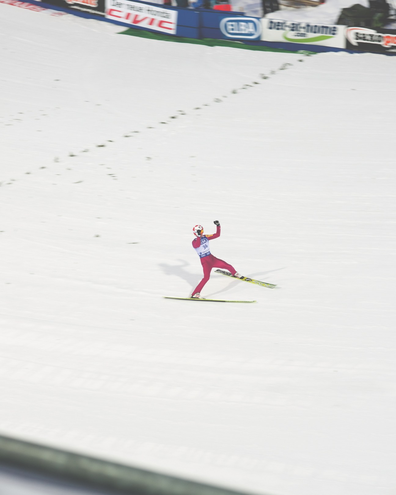 Ski jumper in red suit landing on snowy slope with advertising boards visible above.