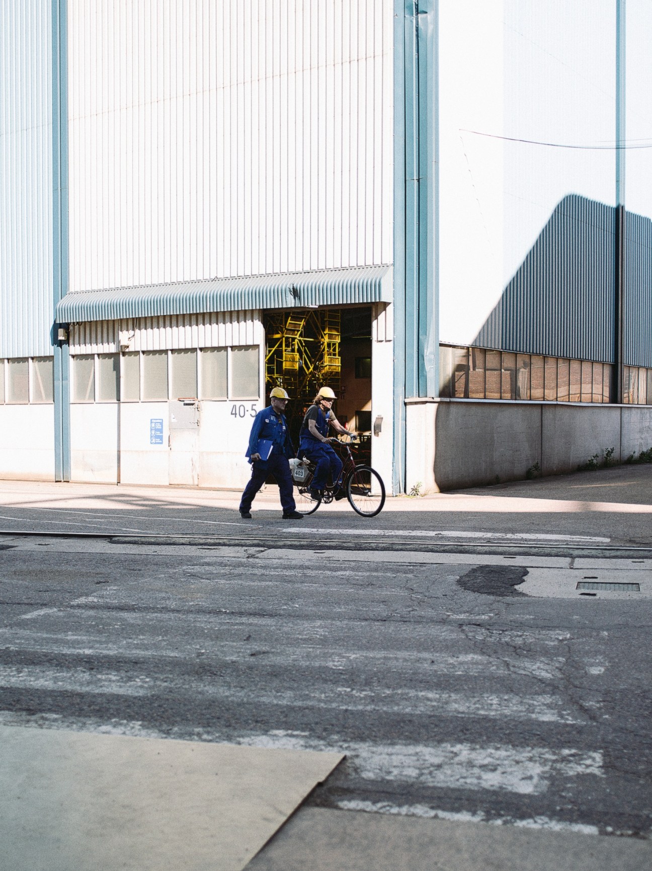 Two workers in blue uniforms and hard hats ride a bicycle past an industrial shipyard building with corrugated metal walls.