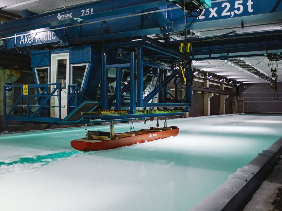 A red-hulled scale model icebreaking cargo ship is suspended in a turquoise ice-filled basin at Aker Arctic's testing facility.