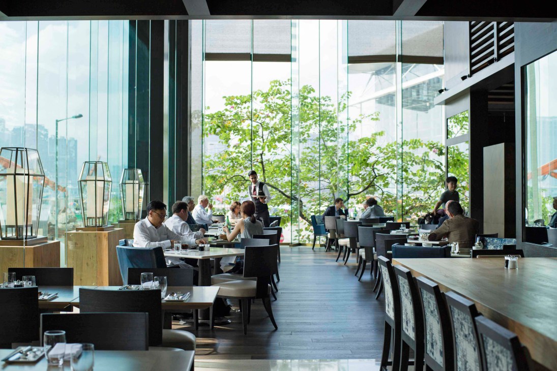 Modern restaurant with floor-to-ceiling windows overlooking Victoria Harbour, featuring contemporary furniture and potted plants.