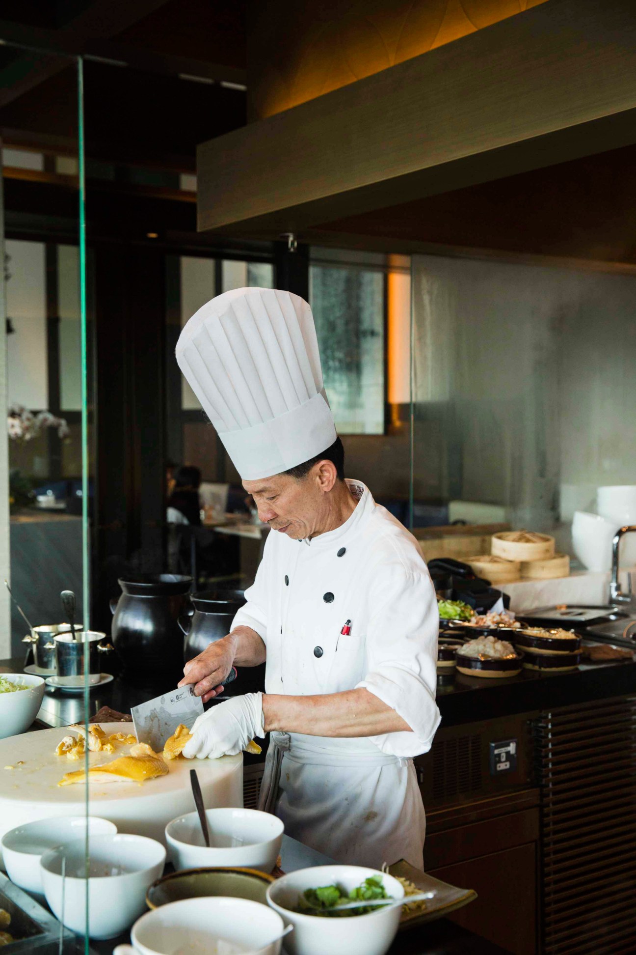 Chef in white uniform and tall chef's hat prepares food at an open kitchen counter.