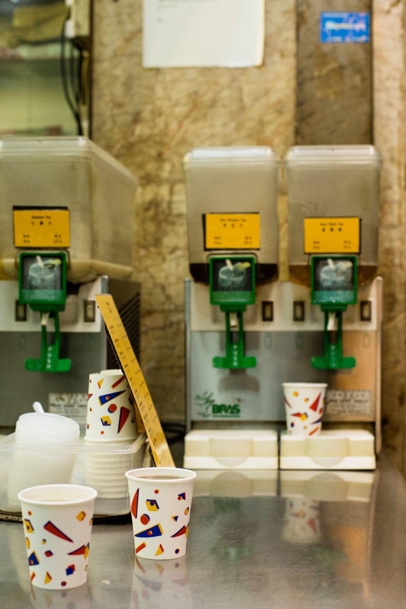 Three white beverage dispensers with yellow and green labels mounted on a cork wall in Kung Lee tea shop.