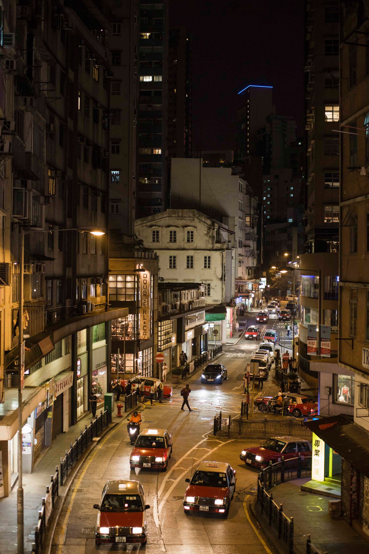 Night view of Hollywood Road in Hong Kong with illuminated storefronts, taxis, and pedestrians on a narrow urban street.