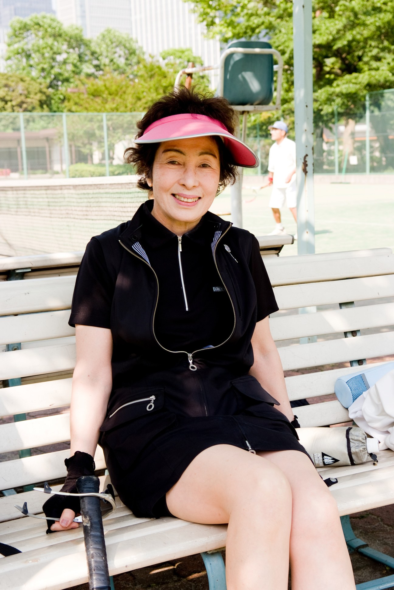 Akio Ohtsuki, 64, sits on a tennis court bench wearing a pink visor and black athletic jacket.