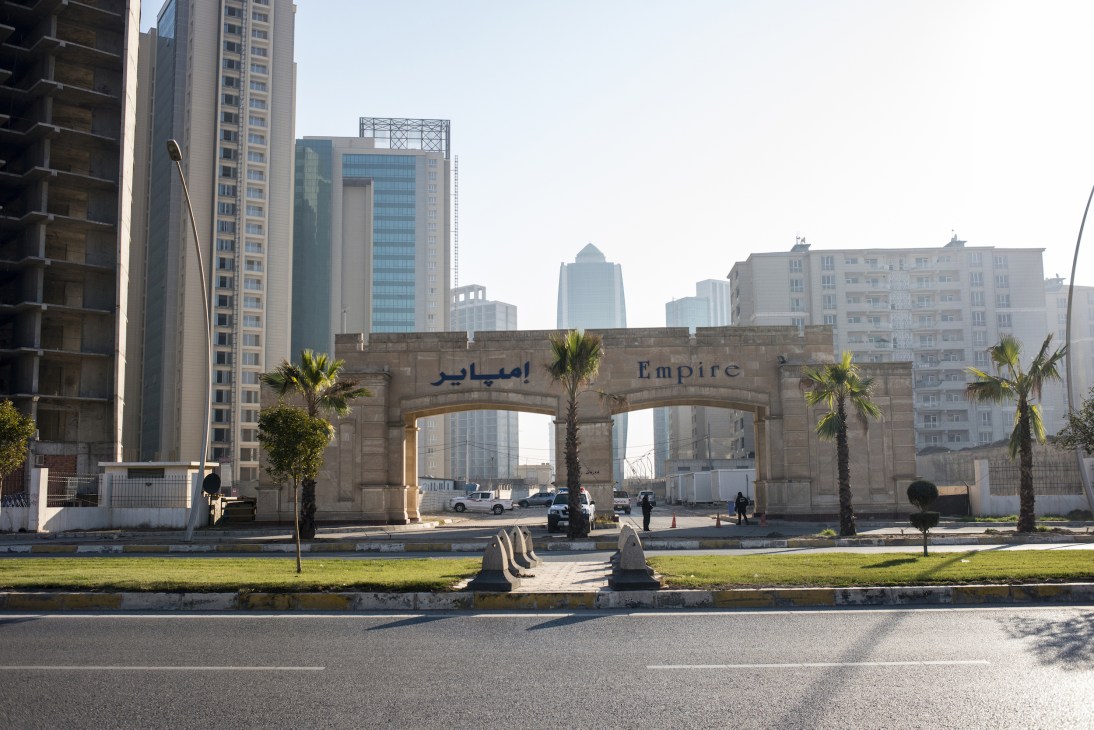 Stone gateway with 'Empire' text and palm trees, with modern high-rise buildings behind in Erbil