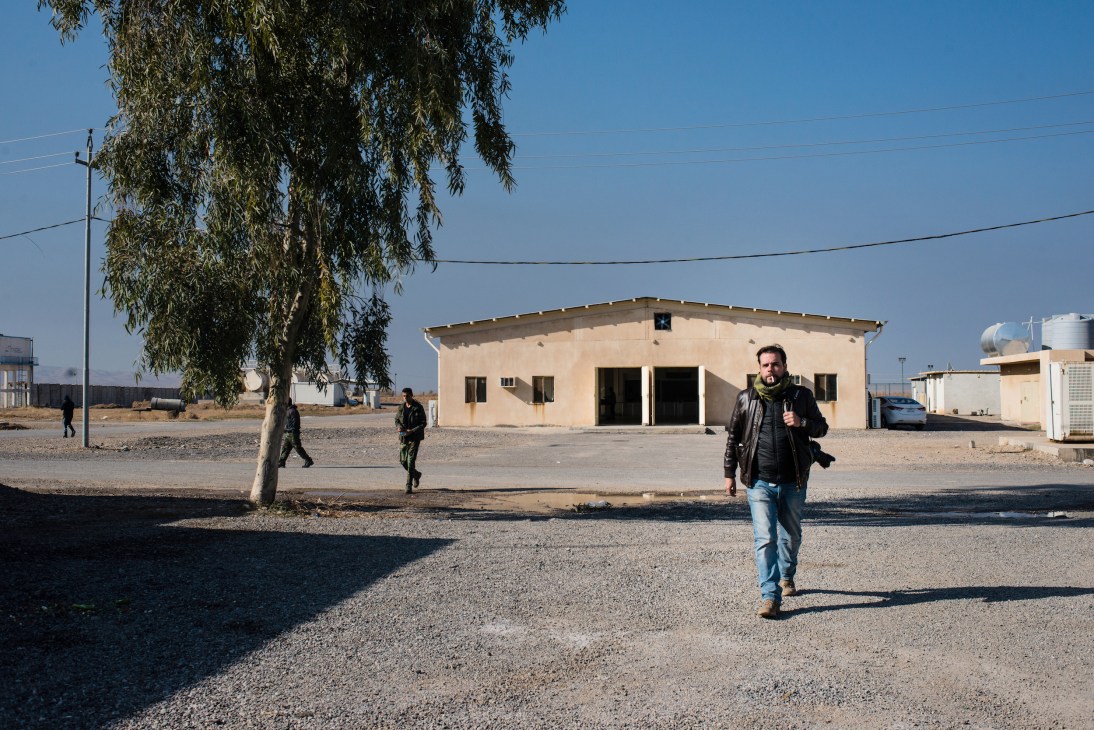 A man in casual clothing stands on a gravel ground near a Peshmerga military base with simple concrete buildings.