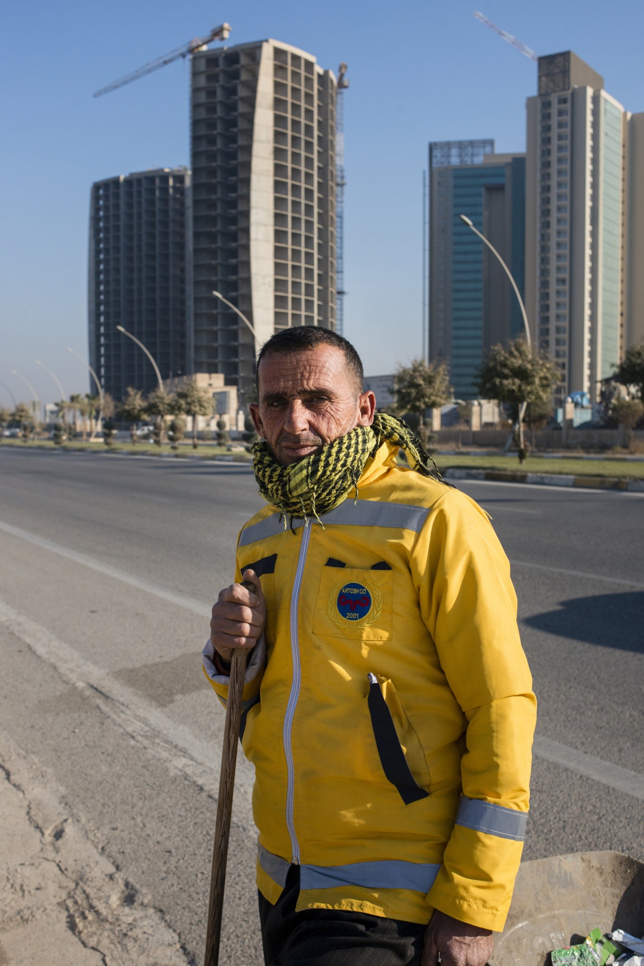Man in yellow jacket and scarf standing on road with modern high-rise buildings under construction behind him.