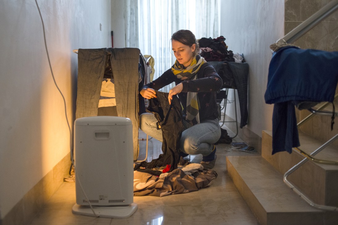 Journalist Oriane Verdier crouches while examining clothing in a sparse room with a heater and hung garments.