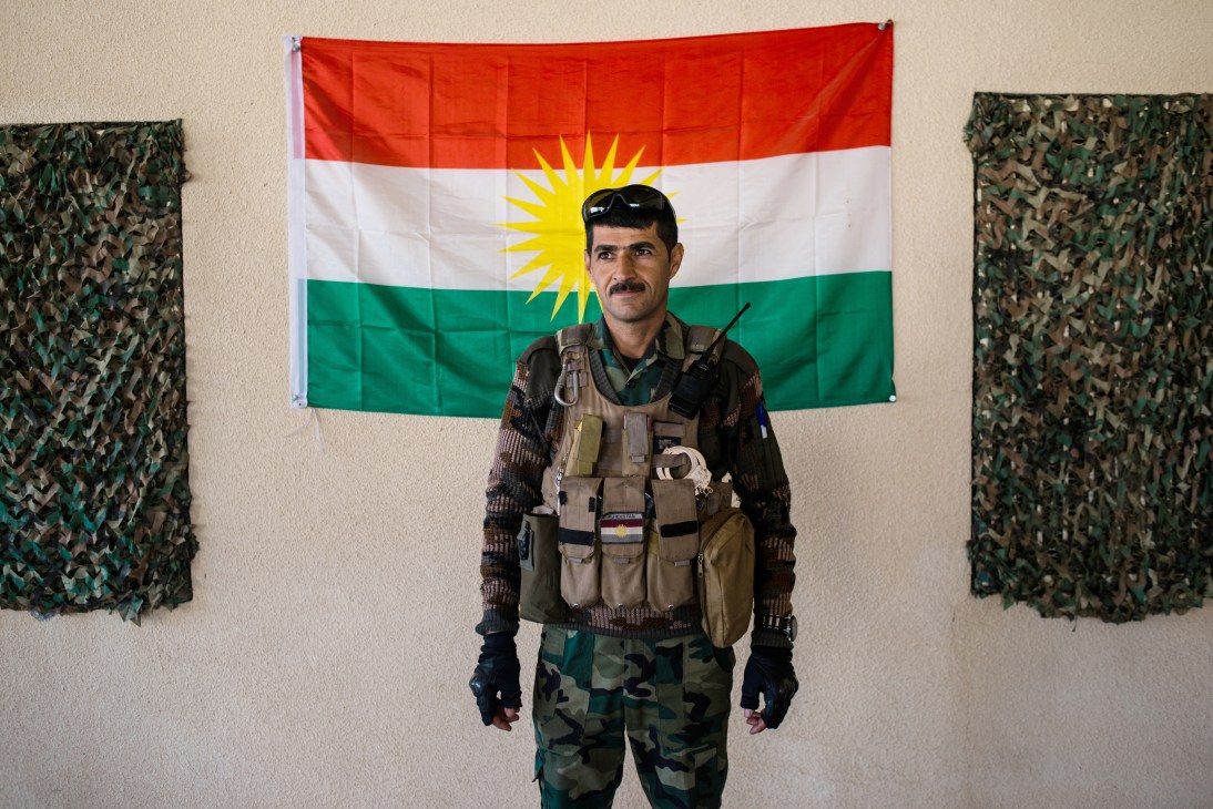 Peshmerga soldier in military gear stands in front of the Kurdish flag at a base near Makhmour.