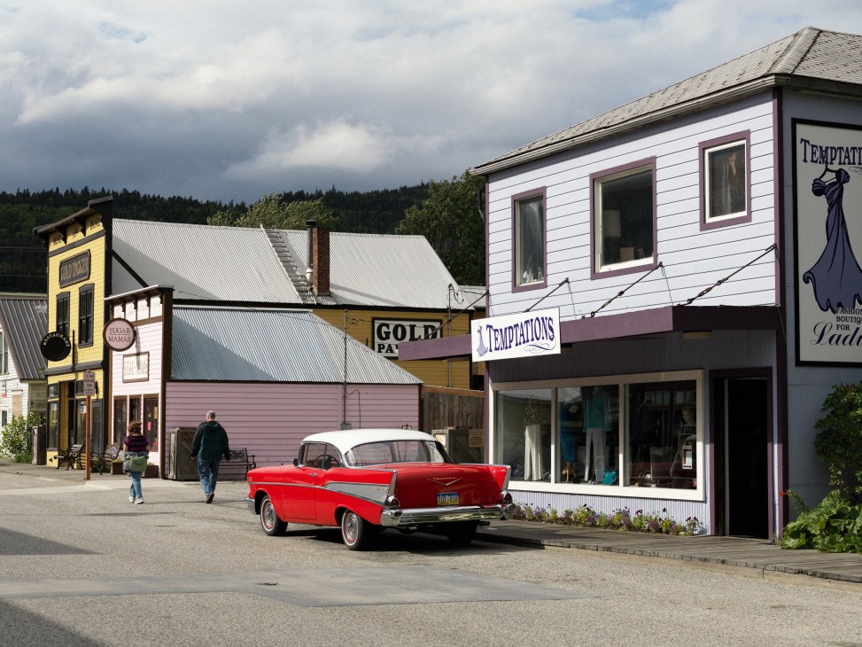 A red 1950s car parked on a street lined with historic Old West-style wooden buildings in Skagway, Alaska.