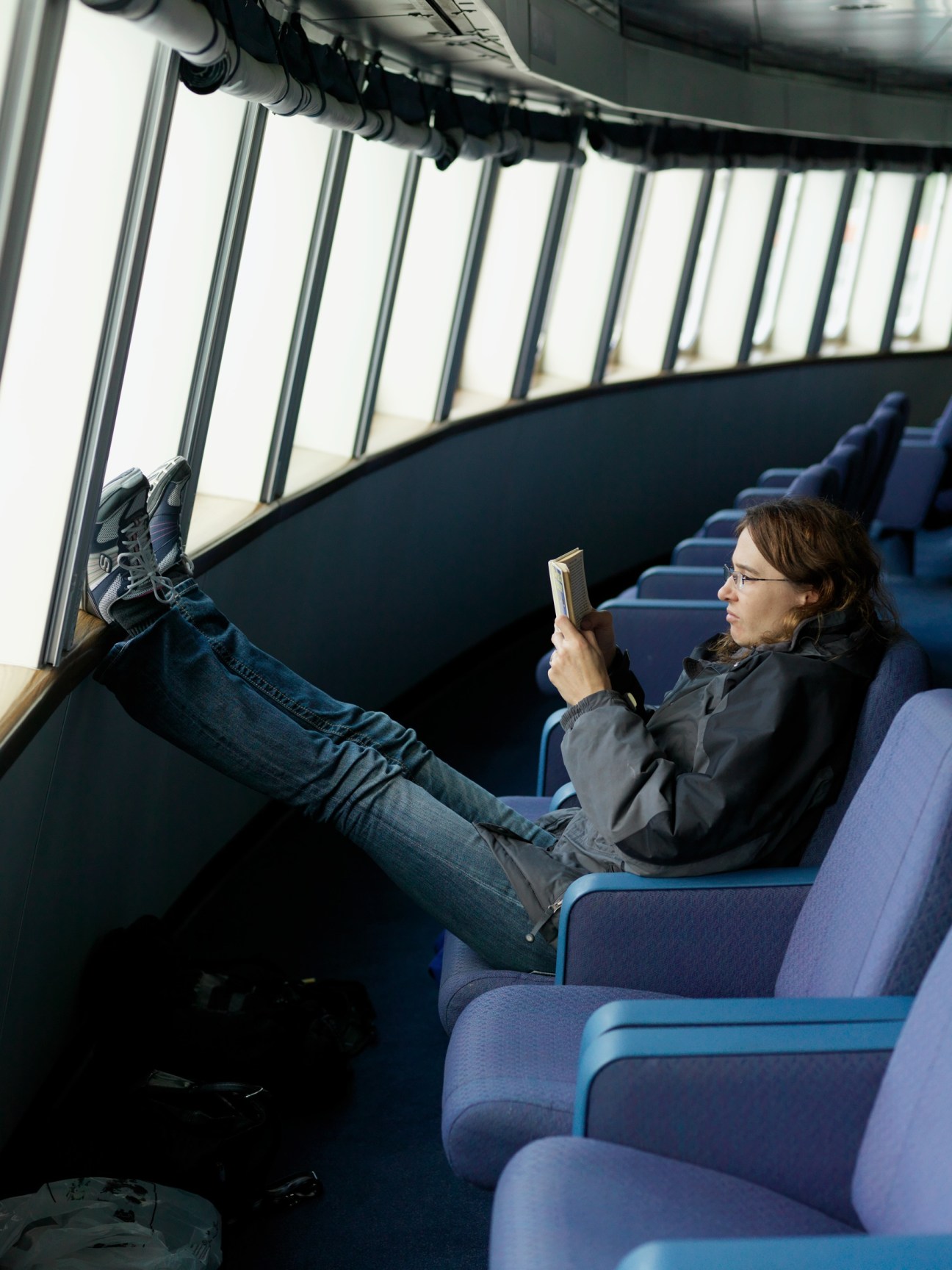Passenger reading in ferry lounge with feet up on window sill and large windows showing sky.