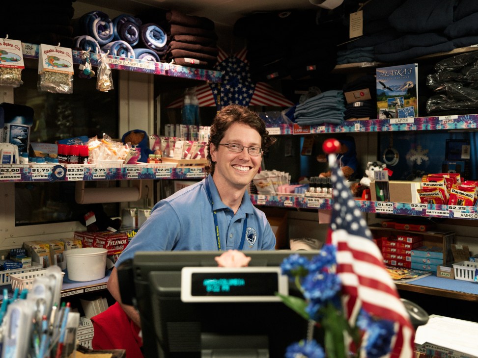 A smiling deck steward stands behind the counter in the M/V Columbia's gift shop, surrounded by Alaska souvenirs and merchandise.