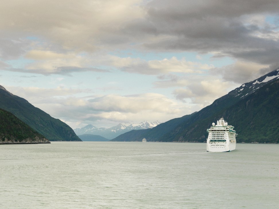 Large white cruise ship sailing through a narrow fjord with snow-capped mountains and forested slopes on either side.