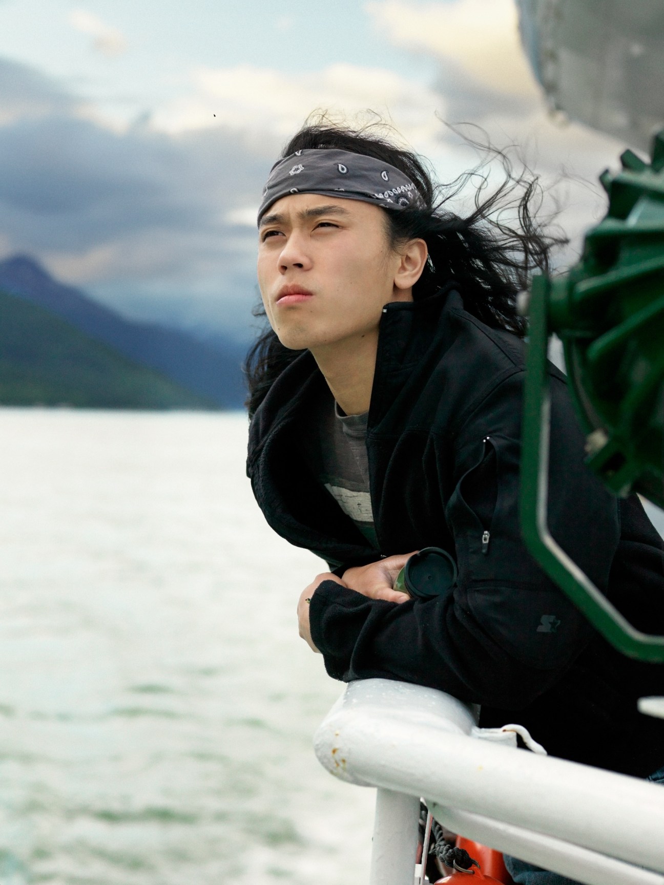 Mitchell Cabrigas leans against the ferry railing with his hair blowing in the wind, mountains visible across the water behind him.