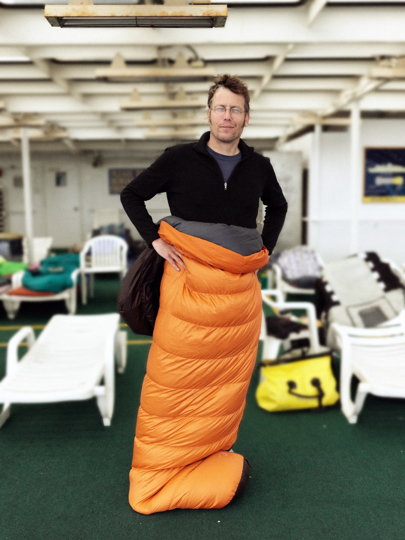 Man in black jacket models bright orange sleeping bag in ferry solarium.