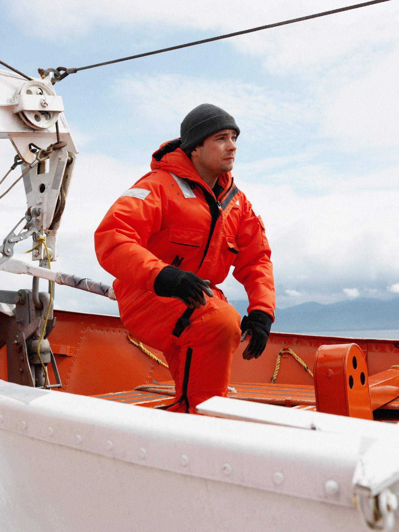 A ferry crew member in a bright orange float suit and gray beanie sits on the deck of the M/V Columbia during a safety drill.