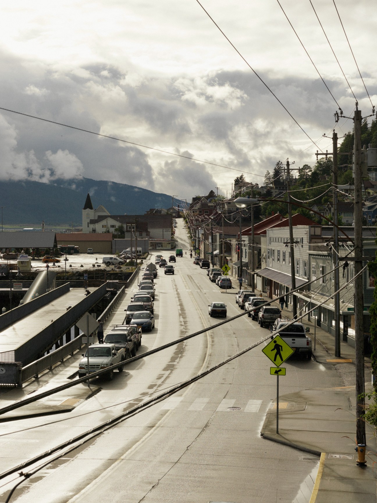 Ketchikan street lined with parked cars, buildings, and mountains under dramatic cloudy sky with golden light breaking through.