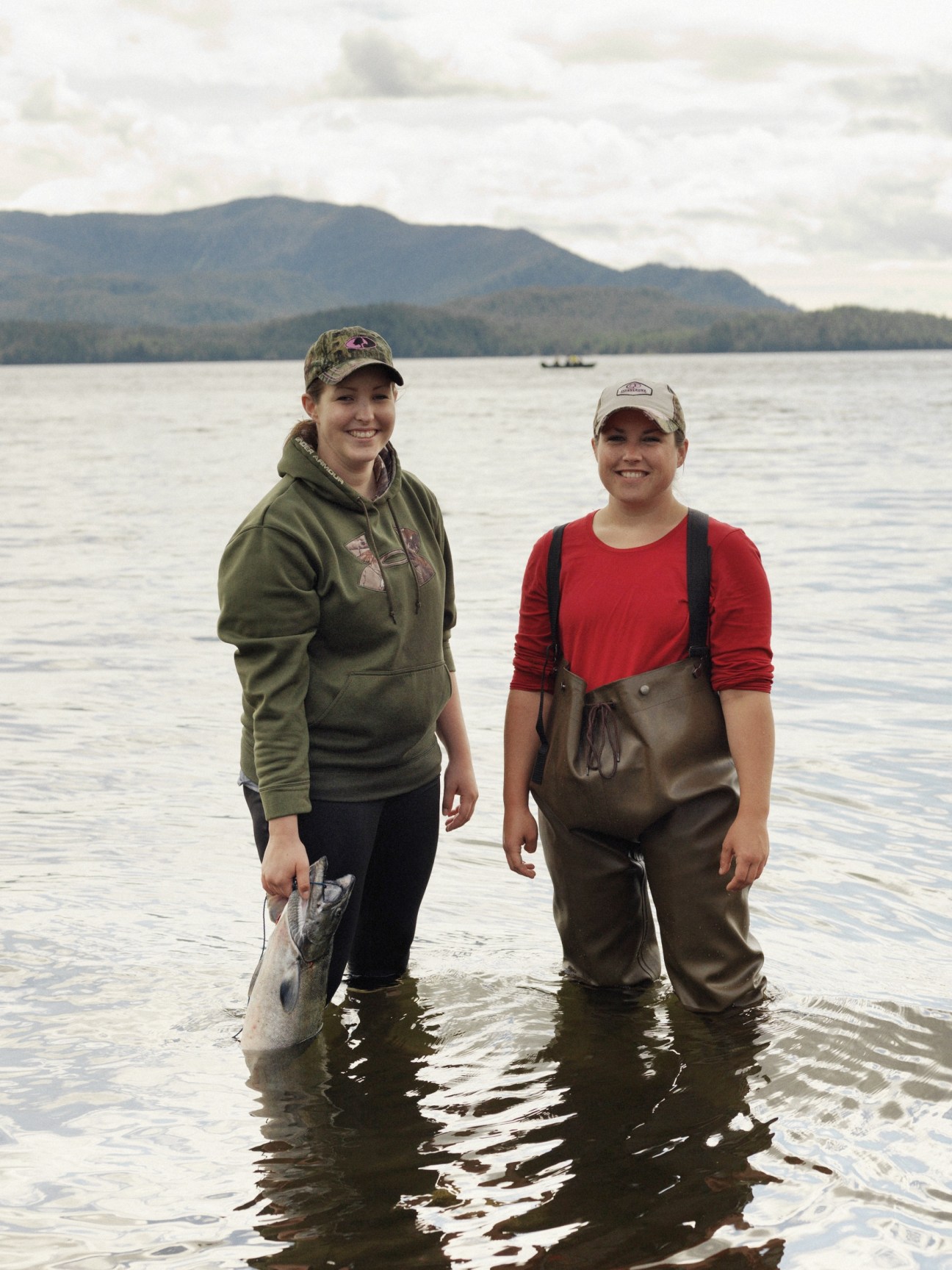 Two women in waders stand in shallow water holding freshly caught salmon.