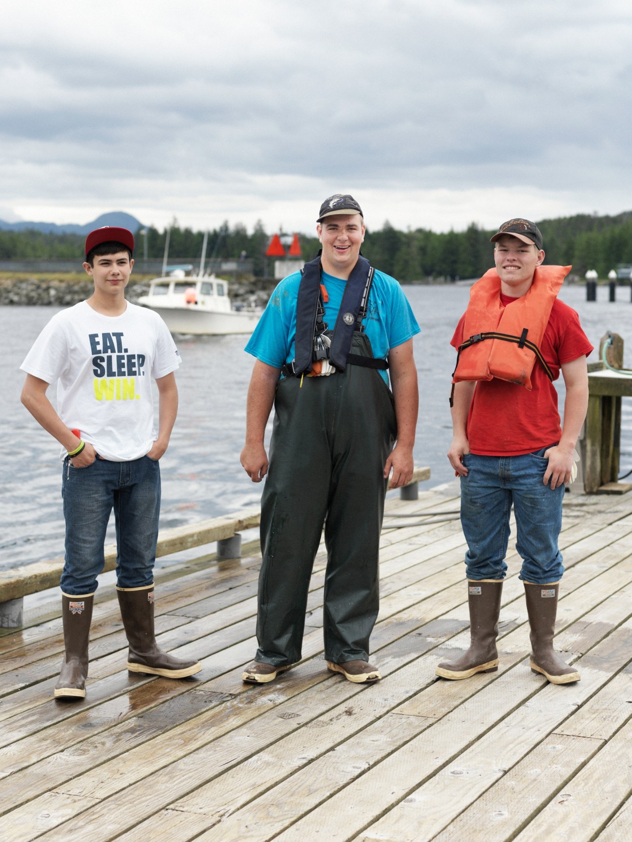 Three young people wearing Xtratuf boots and life jackets stand on a wooden dock with boats and forest in the background.