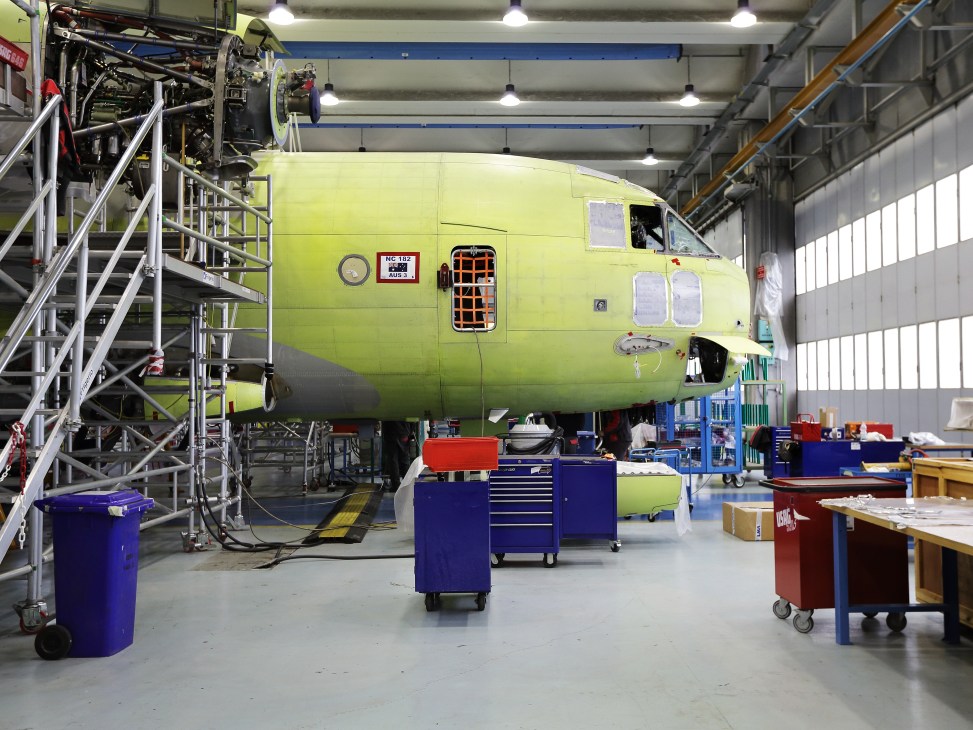 A lime-green C-27J Spartan aircraft under assembly in a factory hangar with blue toolboxes and equipment nearby.