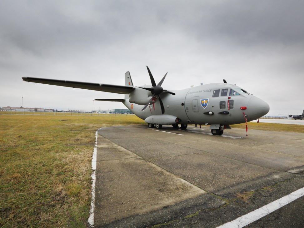 A C-27J Spartan tactical airlifter aircraft parked on an airport apron with grass fields beyond.