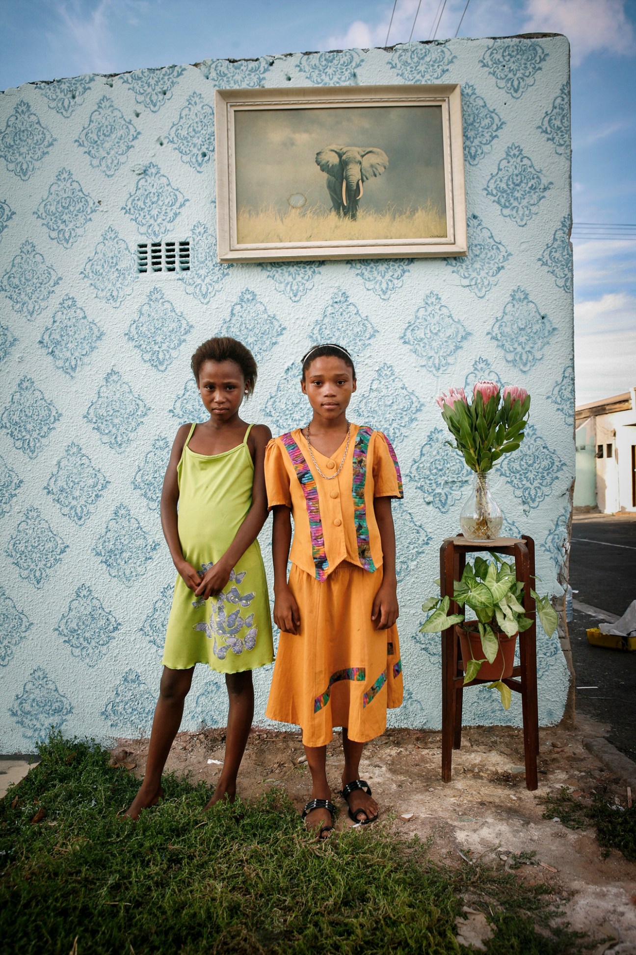 Two girls posing against a blue patterned wall in Woodstock, Cape Town, with a framed elephant painting above and potted flowers beside them.