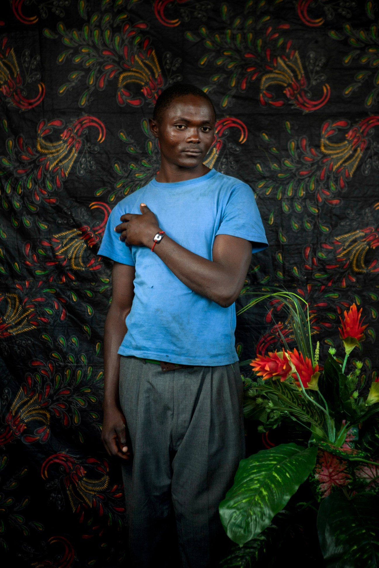 Portrait of Charle Kahalo in a blue t-shirt standing against a patterned dark backdrop with red flowers.