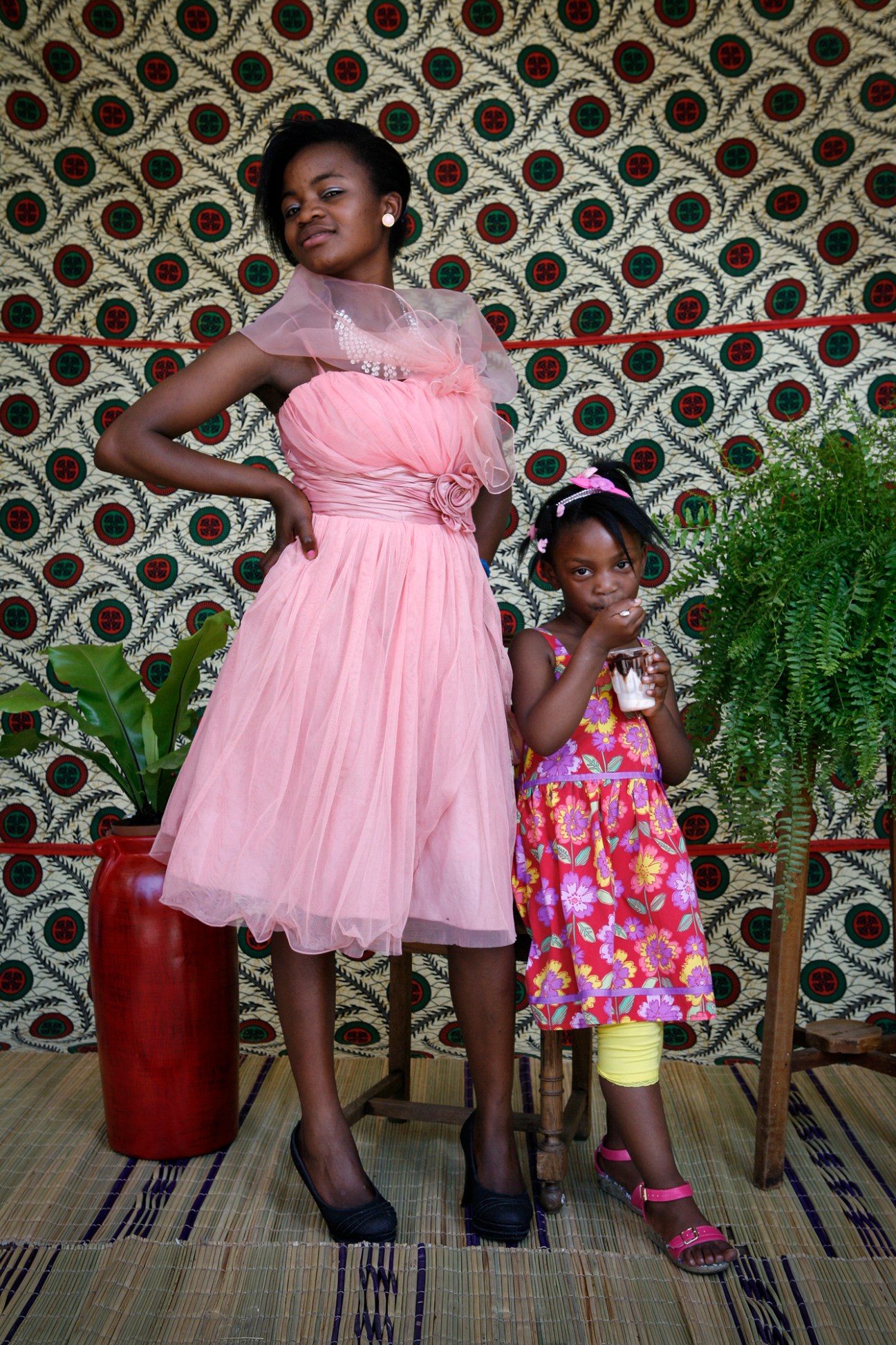 Woman in pink dress and young girl in colorful floral dress pose against patterned backdrop with potted plants in makeshift studio