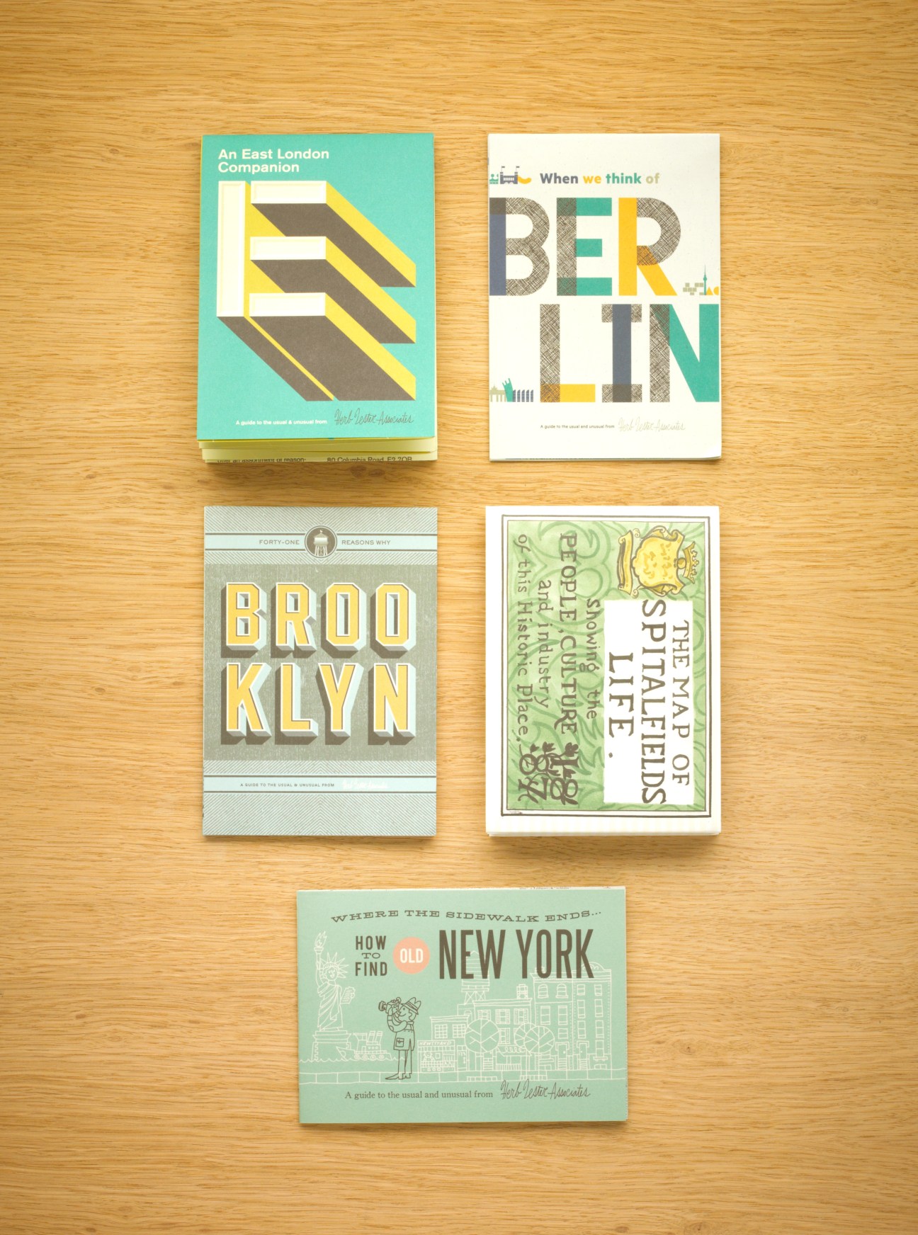 Five colorful Herb Lester guide maps displayed on a wooden surface, including covers for East London, Berlin, Brooklyn, and New York.