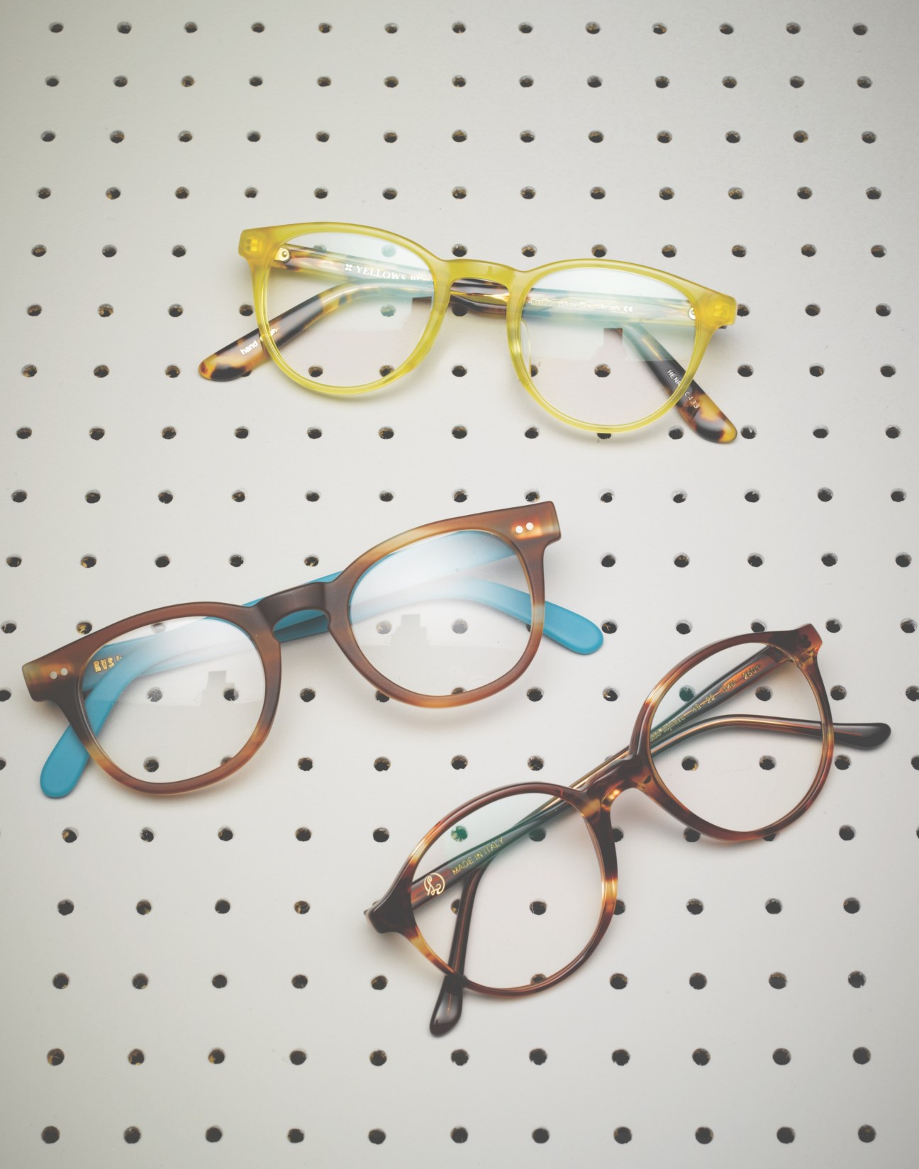 Four handmade eyeglass frames displayed on a perforated white background: a yellow tortoiseshell pair at top, a brown pair with blue temples, and two tortoiseshell pairs below.