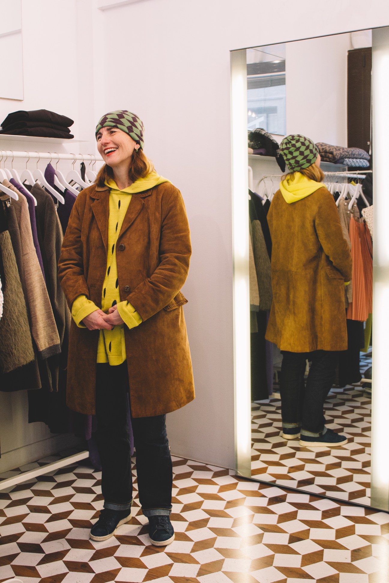 Woman in brown coat and yellow scarf smiles in Almira Sadar boutique with geometric floor and clothing rack.