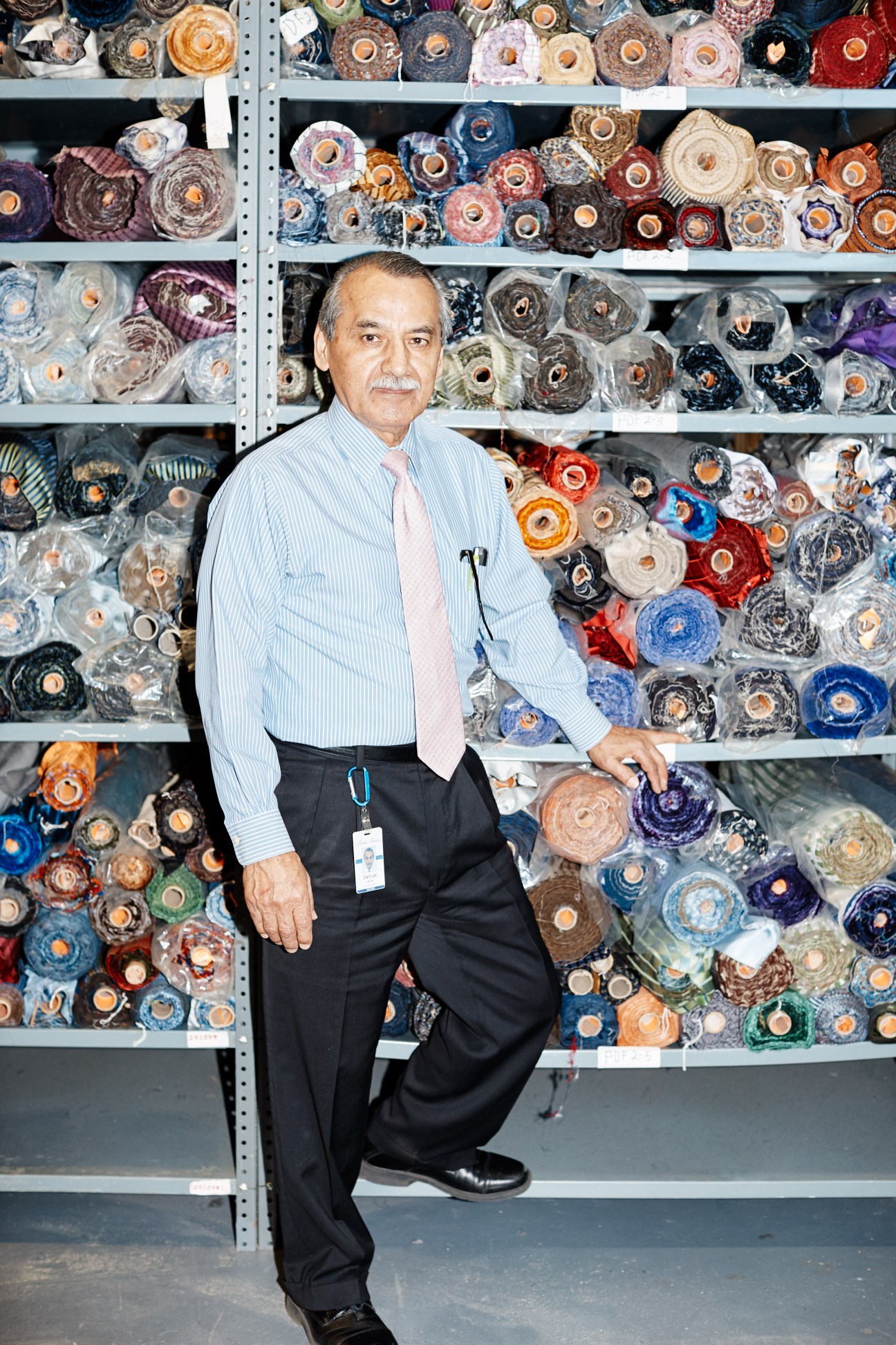 Man in business attire standing in front of shelves of colorful fabric bolts and textile materials.