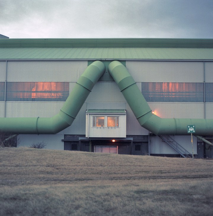 Industrial aluminum plant with large green pipes forming an arch over a corrugated metal building.