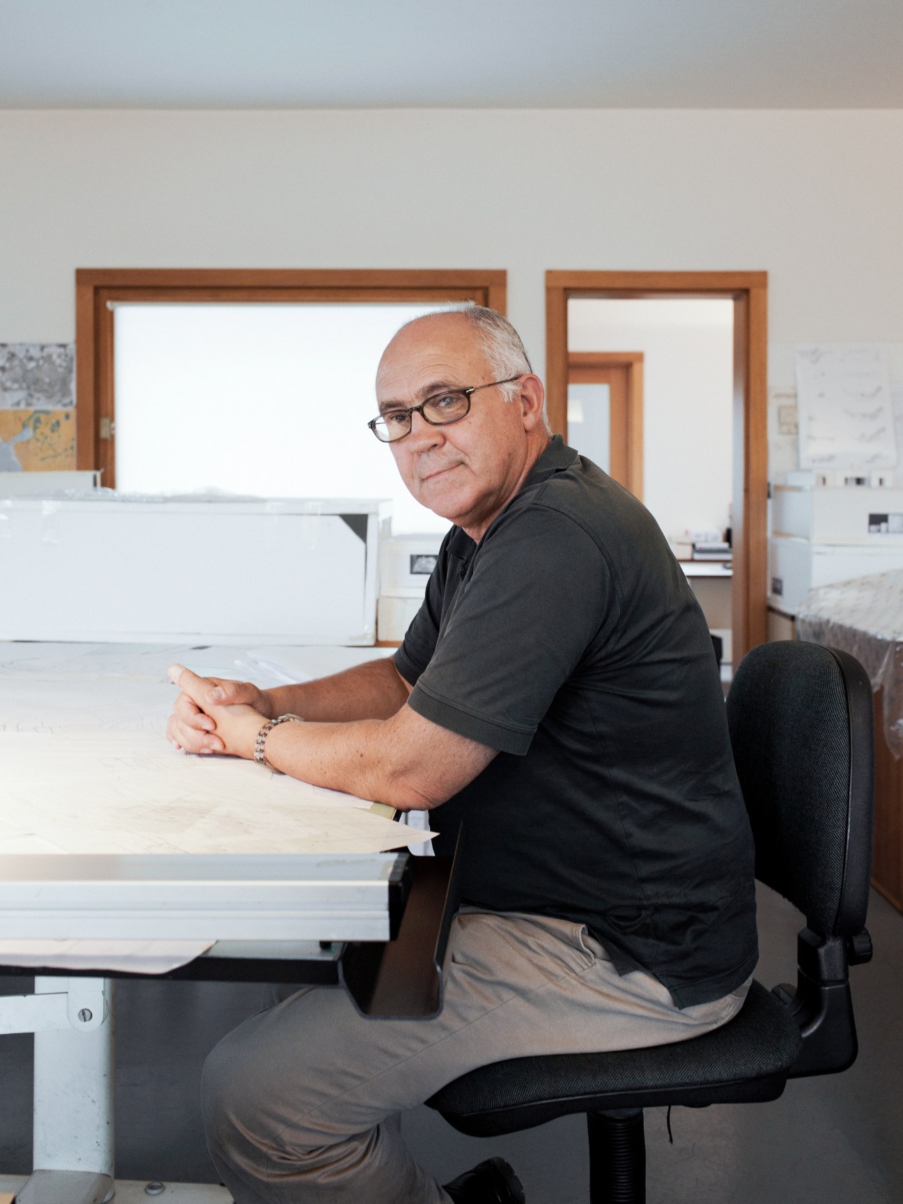 A man wearing glasses and a black shirt sits at a desk with architectural drawings in Siza's office.