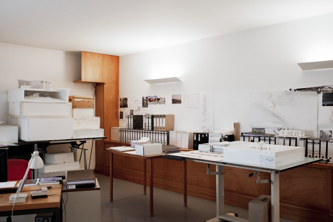 Álvaro Siza's modernist office interior with wooden work tables, architectural models, drawings, and storage displayed on white walls.