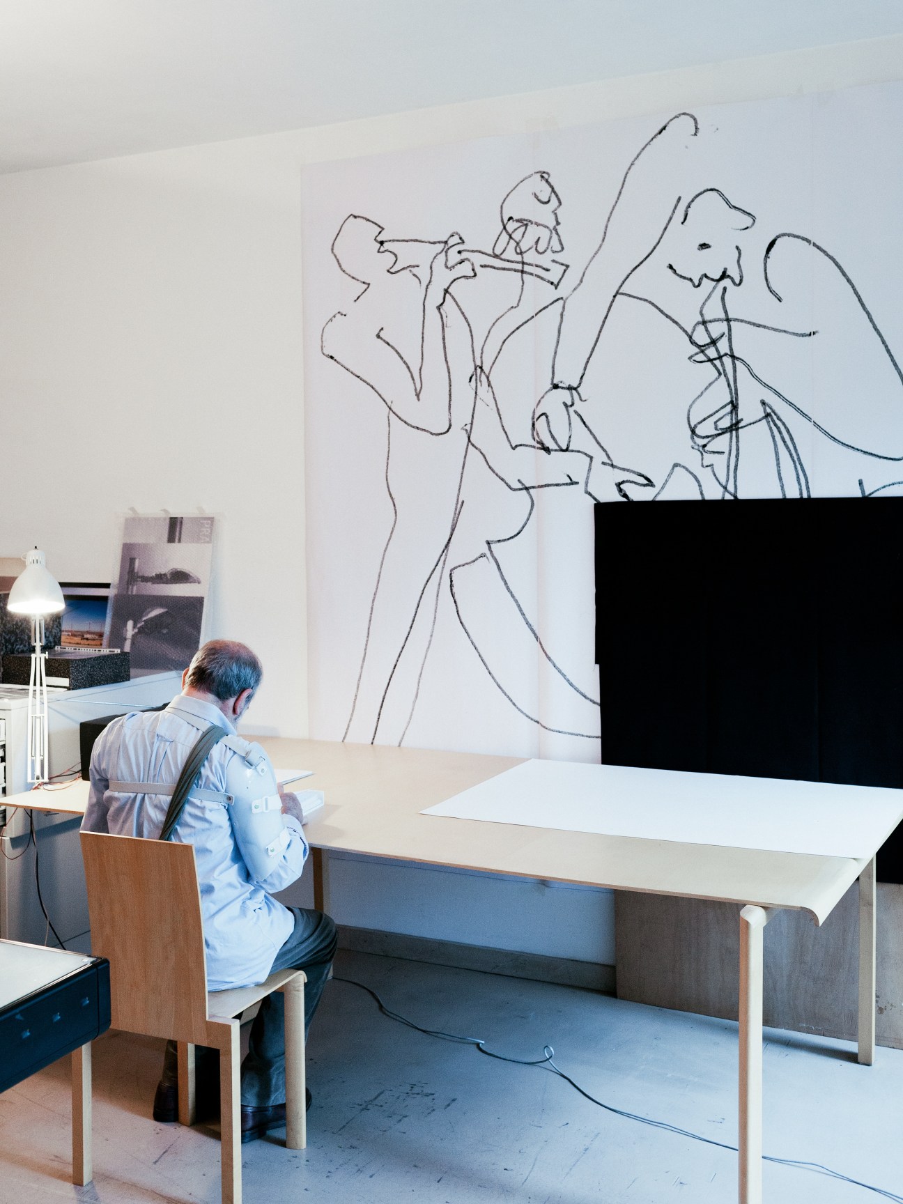 Álvaro Siza seated at a desk in his Porto office, working beneath a large line drawing on the wall.